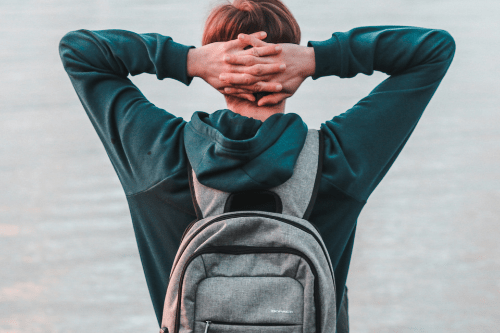 Boy wearing backpack looking out at the water with hands clasped behind his head